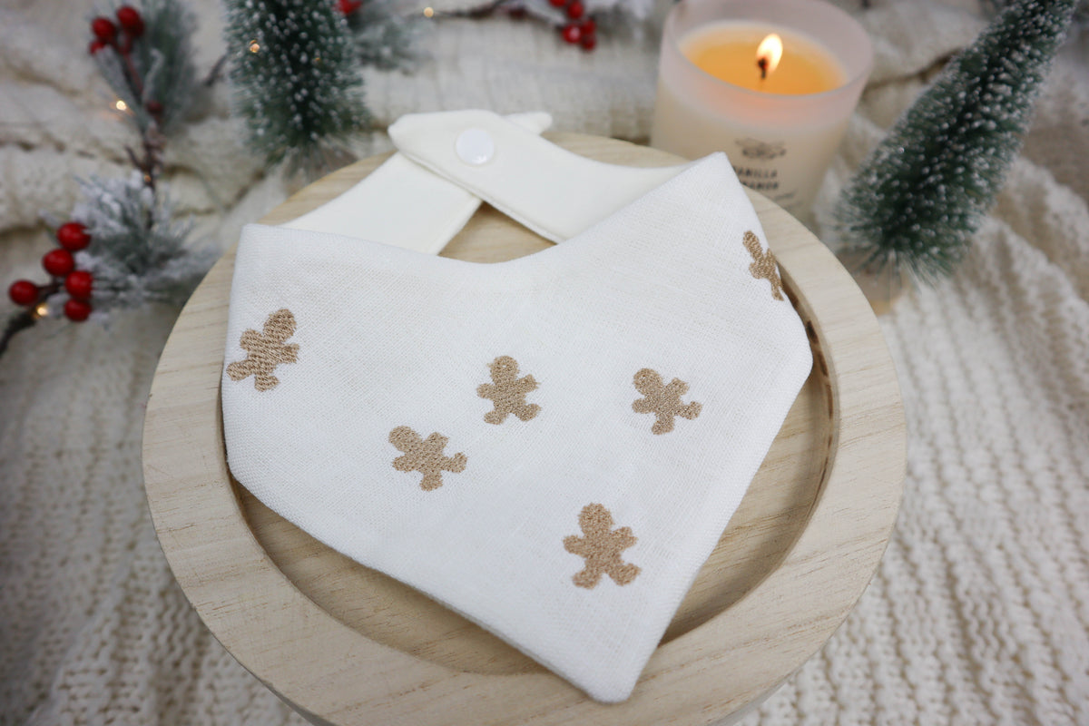 White dog bandana with tan gingerbread man designs on a wooden plate with a candle and Christmas decorations in the background.