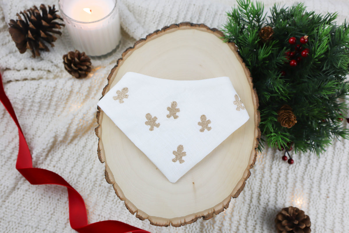 Wooden coaster with a white dog bandana featuring gingerbread man designs, surrounded by Christmas decorations.