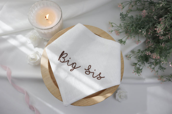 White dog bandana with 'Big Sis' embroidery on a wooden board with a candle and flowers in the background.