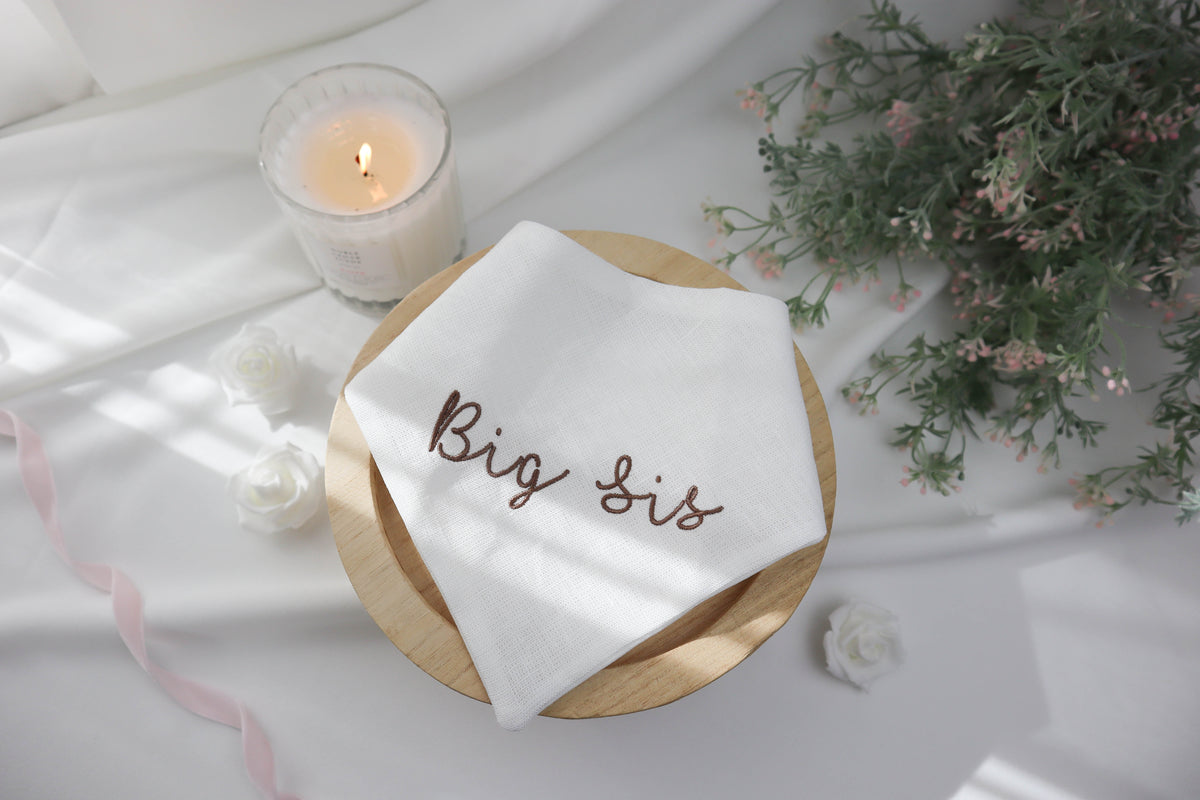 White pet bandana with 'Big Sis' embroidery on a wooden plate with a candle and flowers in the background.