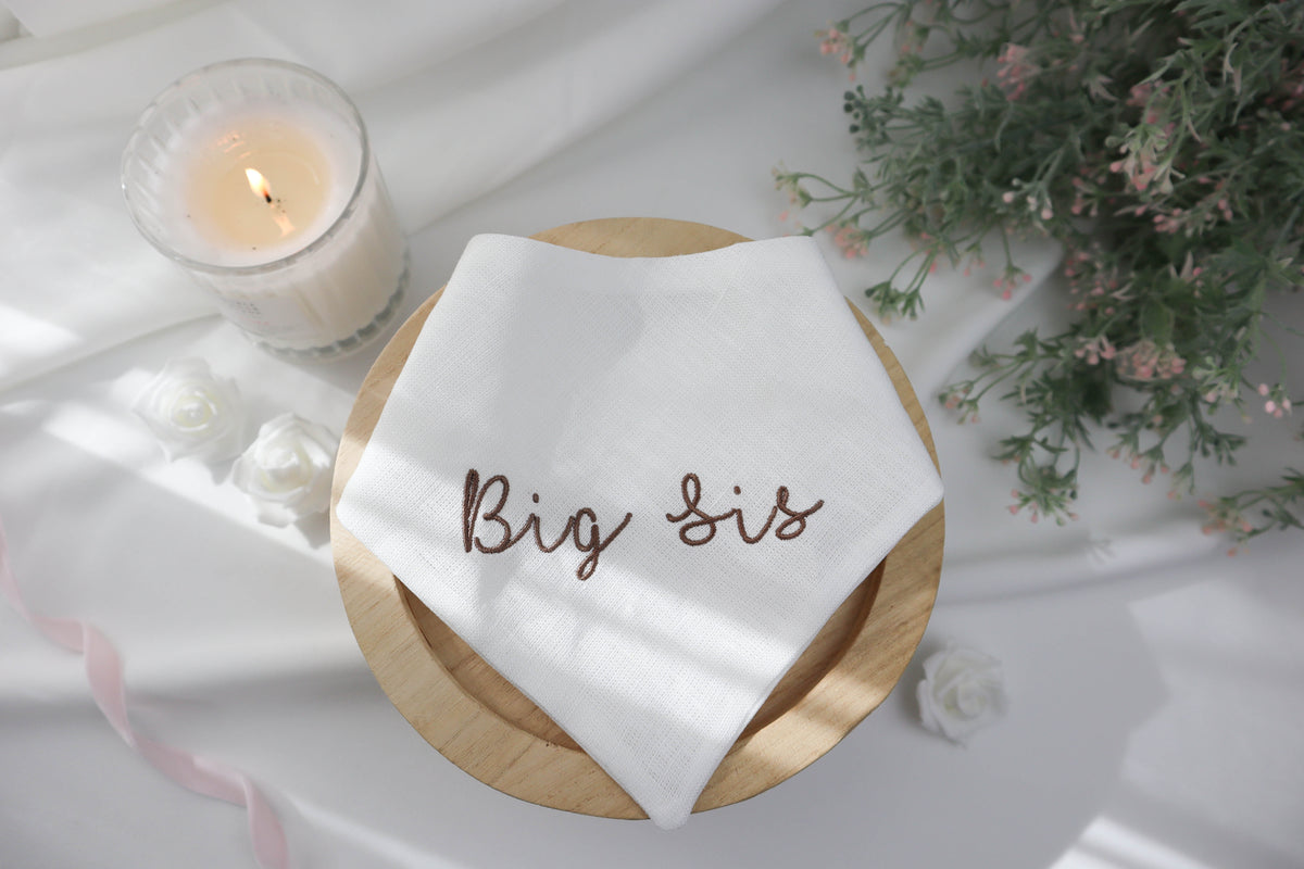 White pet neckerchief with 'Big Sis' embroidery on a wooden board, surrounded by a candle and flowers.