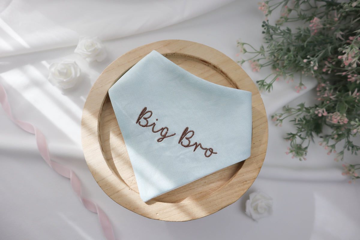 Light blue baby bandana with 'Big Bro' embroidery on a wooden plate with flowers in the background