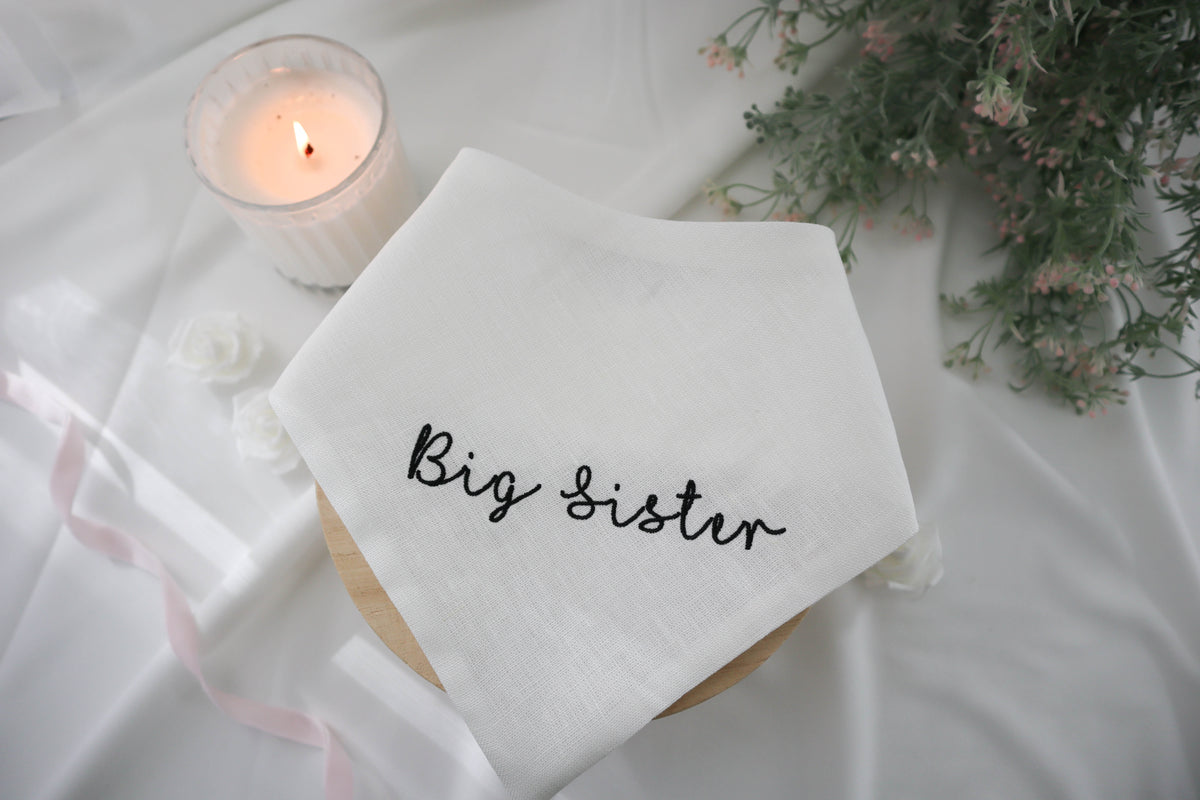 White dog bandana with 'Big Sister' text, lit candle, and flowers on a white surface