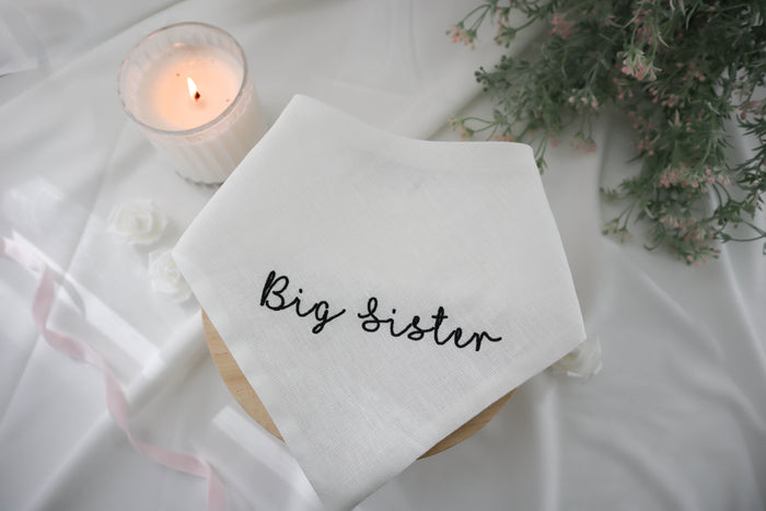 White dog bandana with 'Big Sister' text, lit candle, and flowers on a white surface