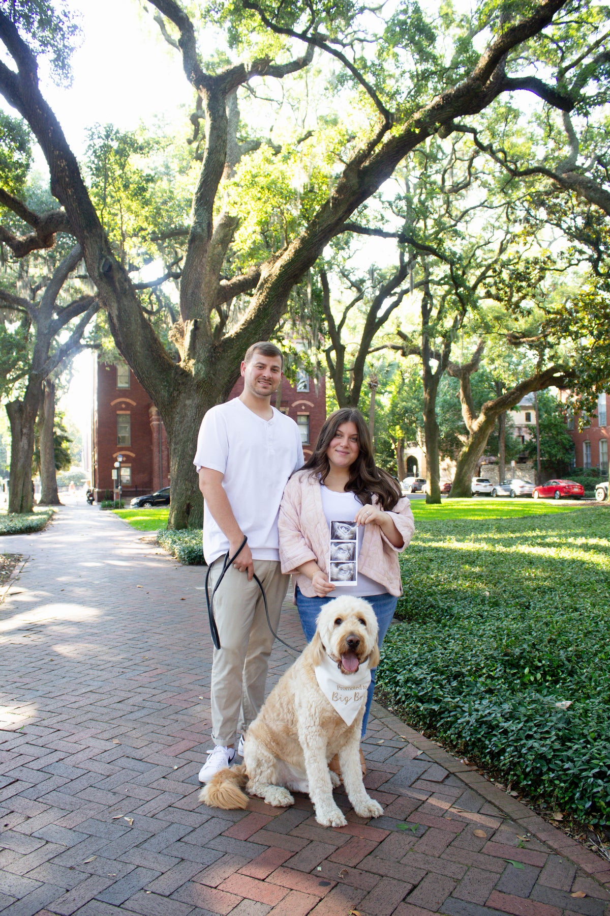 Man and woman with a dog wearing a big bro dog bandana on a leash in a park-like setting with trees and buildings in the background.