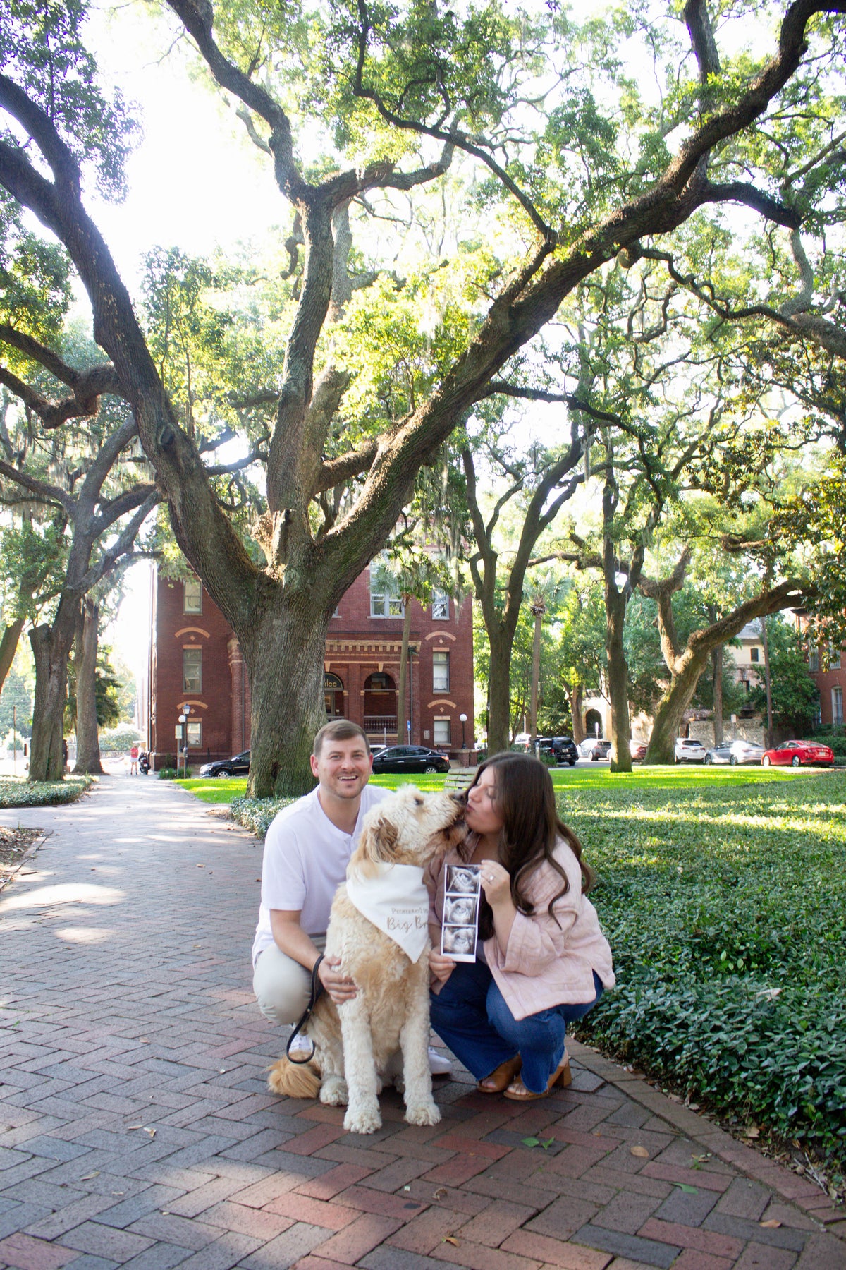 Couple with a dog wearing a big bro dog bandana on a brick path in a park-like setting with trees and buildings in the background.