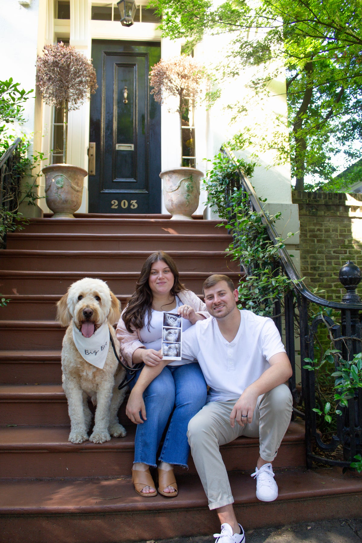 Two people and a dog  wearing a dog bandana with big bro on it sitting on steps in front of a house with a blue door.