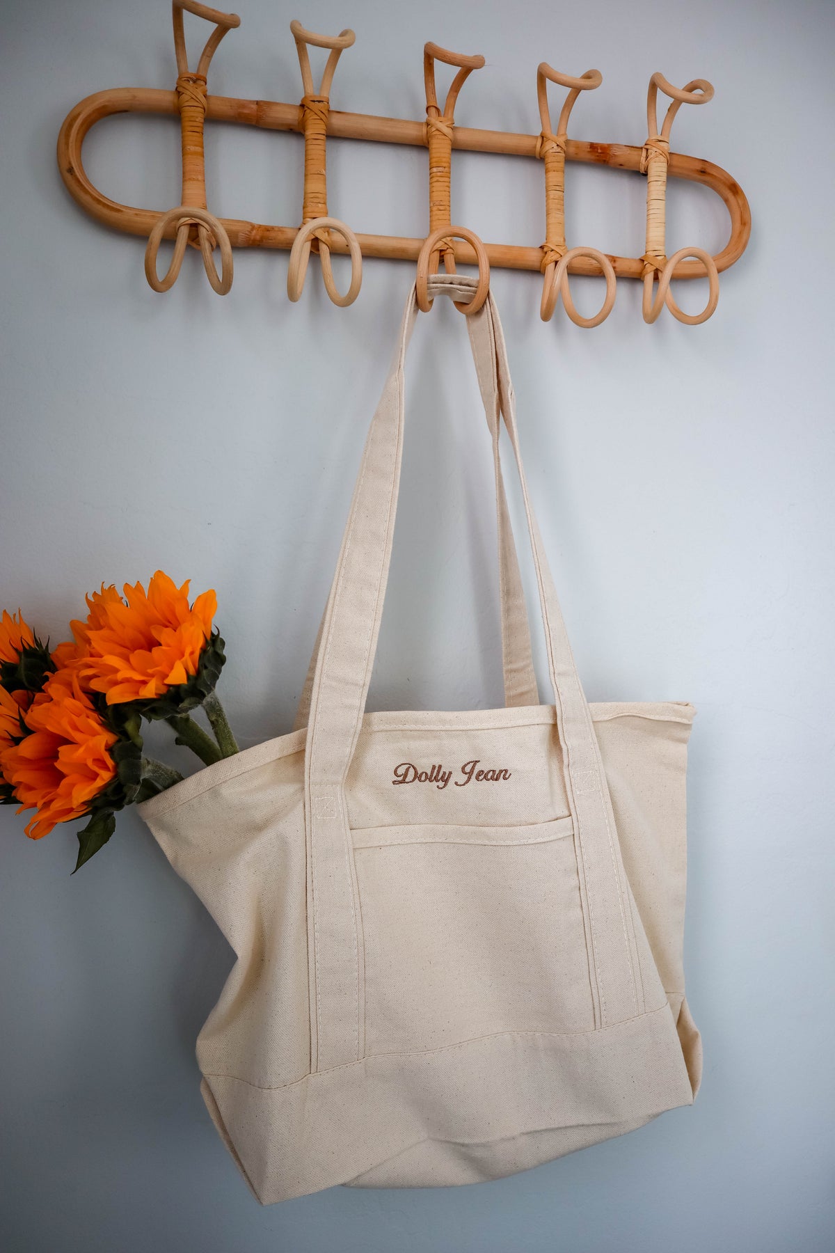 Beige tote bag with 'Dolly Jean' embroidery hanging on a wooden hook against a light blue wall.