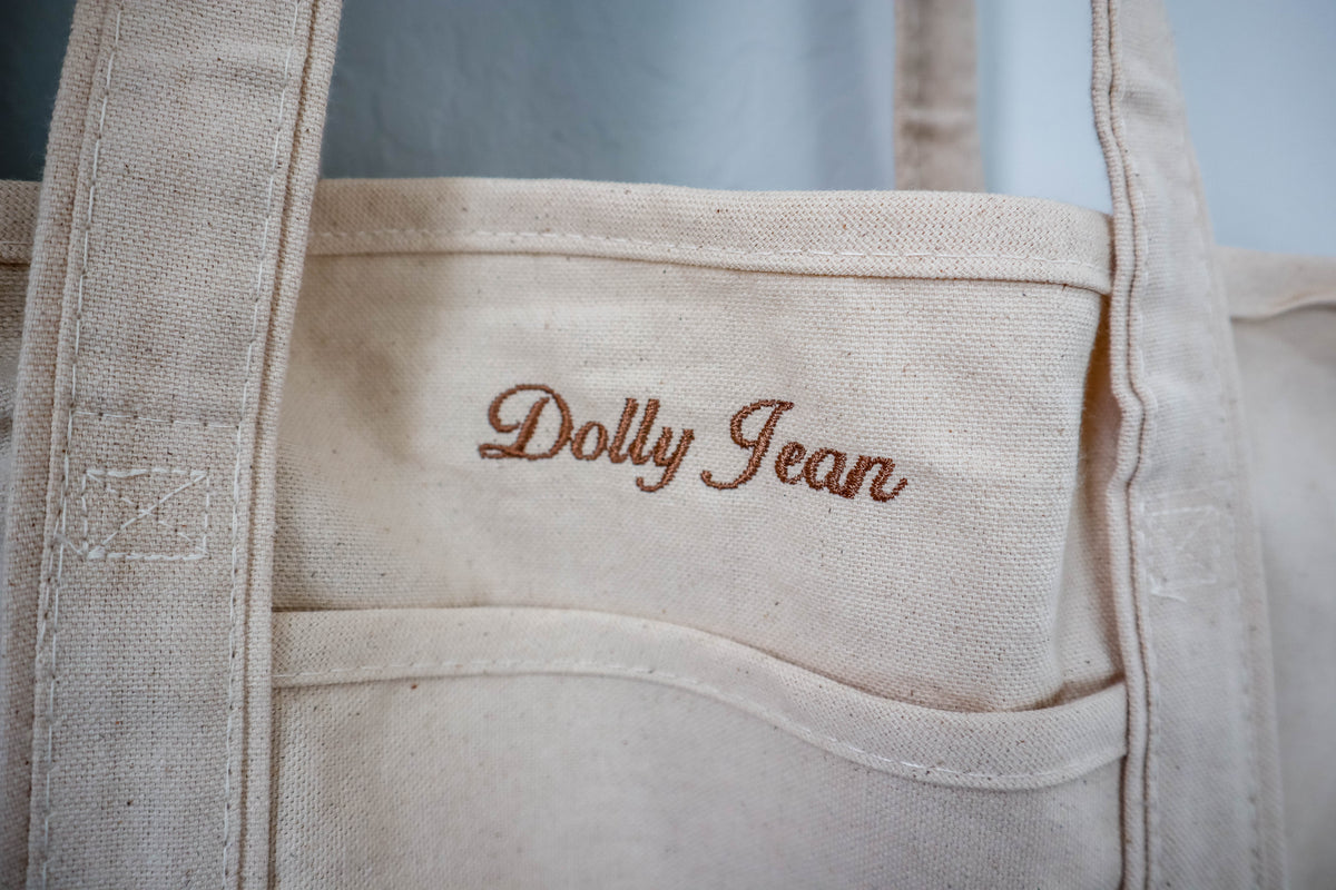 Beige tote bag with 'Dolly Jean' embroidery on a light blue background