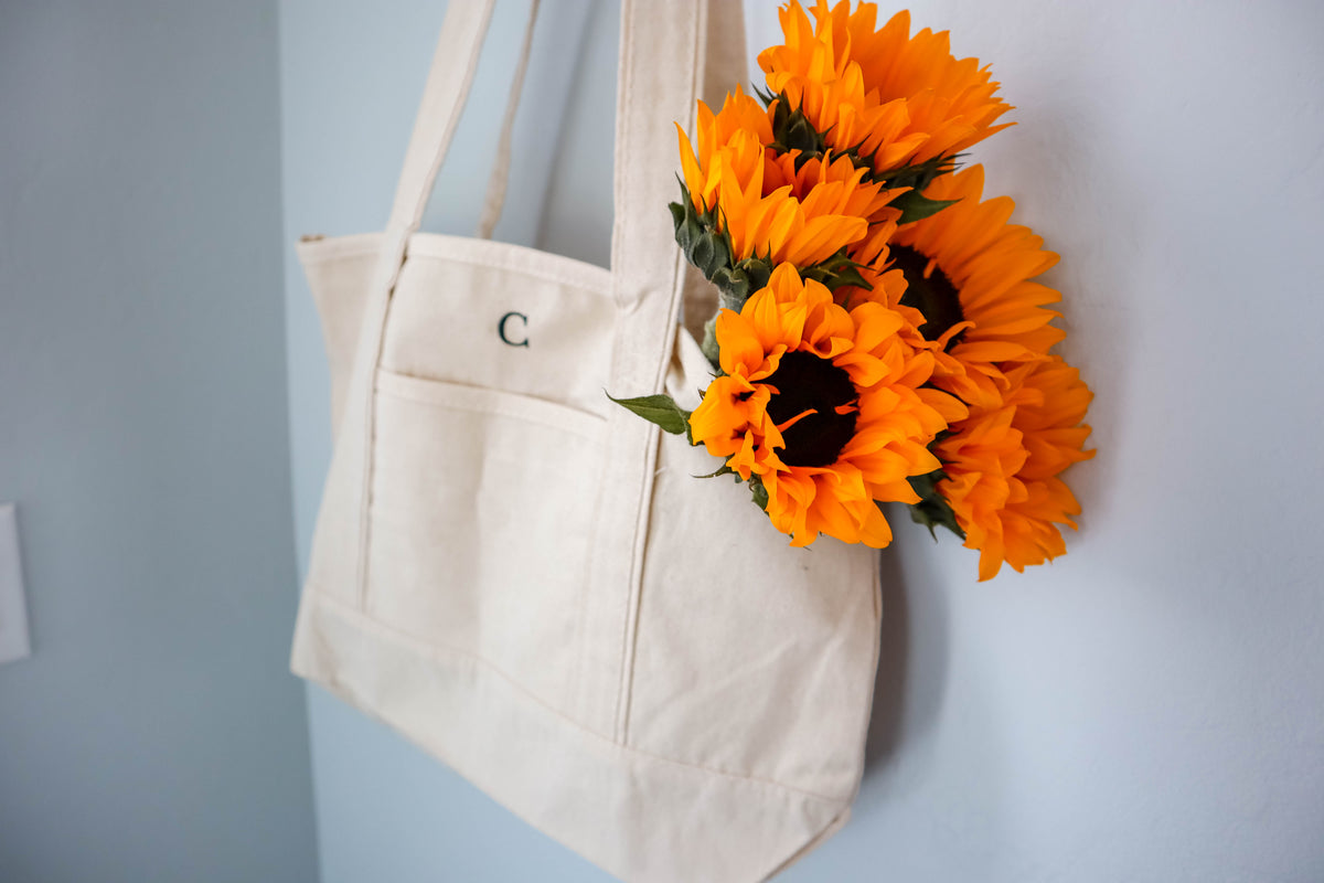 Beige tote bag with a monogram 'C' next to a bouquet of sunflowers on a light blue background