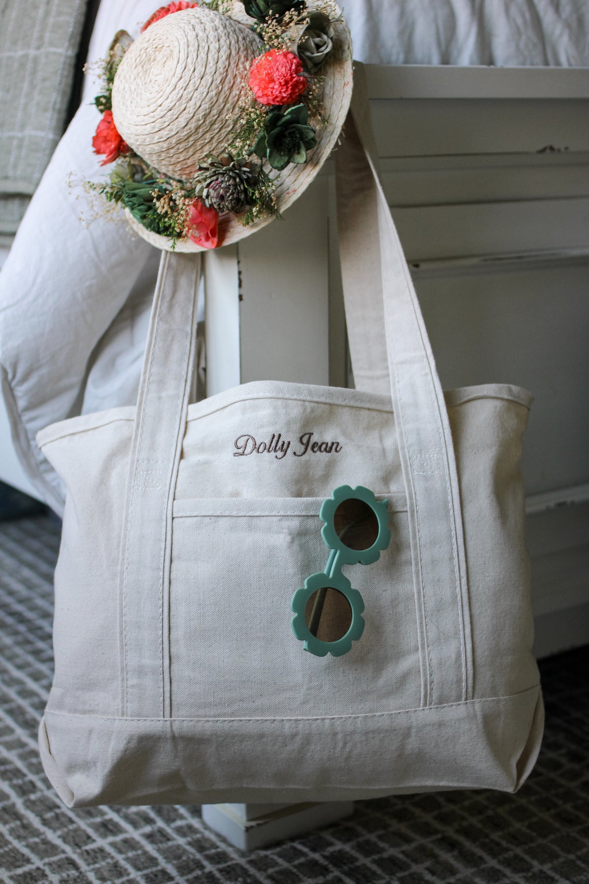 White tote bag with 'Dolly Jean' branding and floral sun hat on a textured surface.