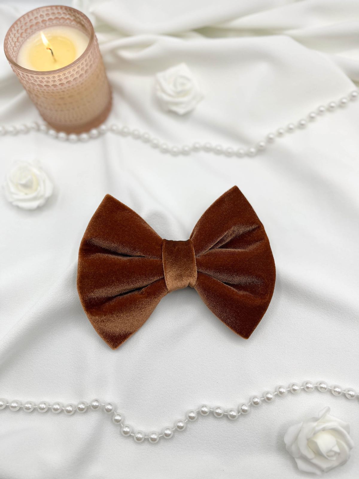 Brown velvet bow tie on a white fabric background with a candle and pearls.