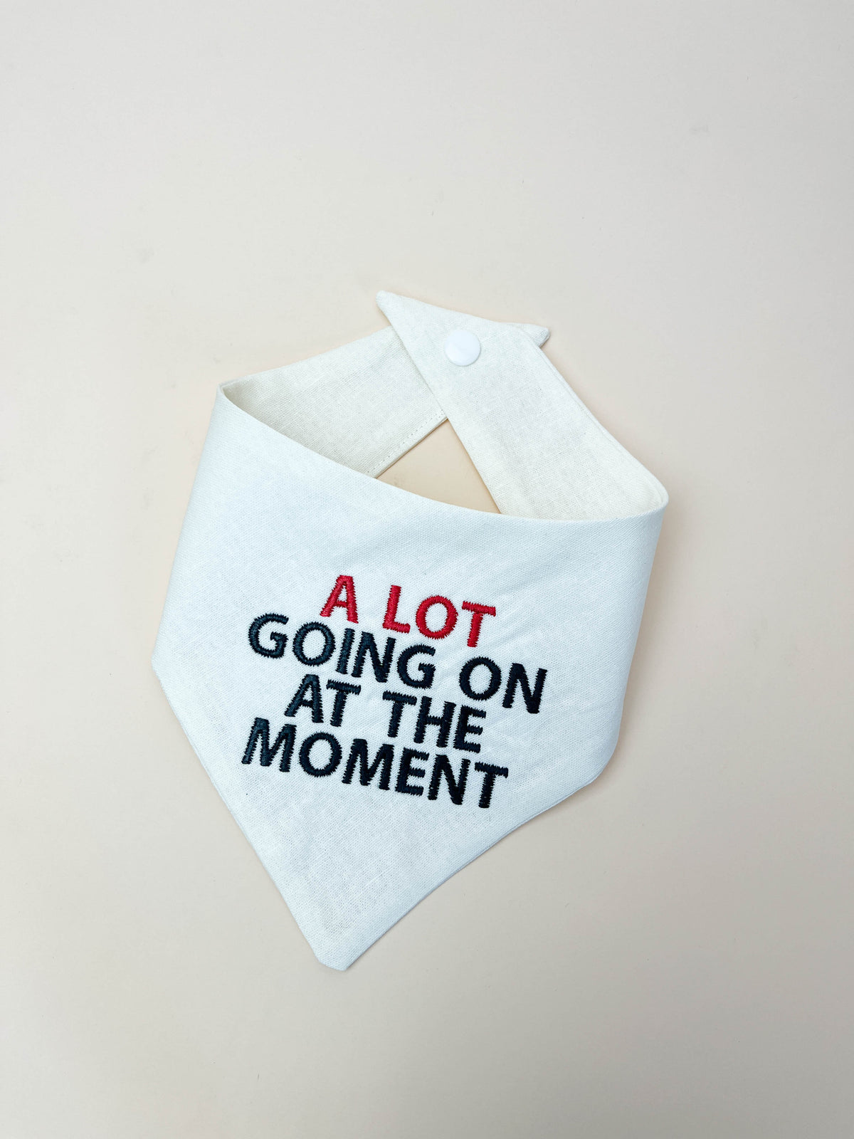 White bandana bib with text 'A Lot Going On At The Moment' on a beige background