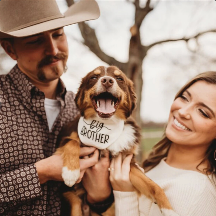 Man and woman holding a dog wearing a 'Big Brother' dog bandana outdoors.