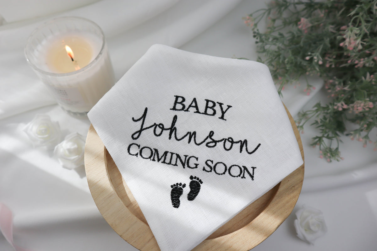 White dog bandana with 'Baby Johnson Coming Soon' text on a wooden plate with a candle and flowers in the background.