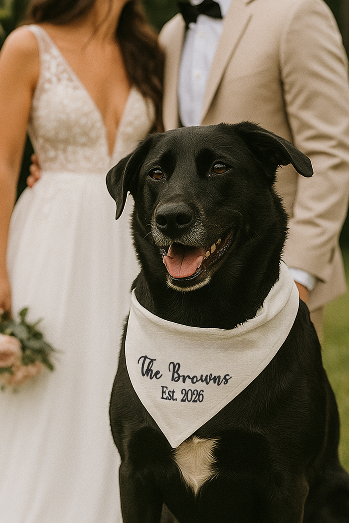 Black dog wearing a bandana with 'The Browns Est. 2026' text, standing in front of a couple in wedding attire.