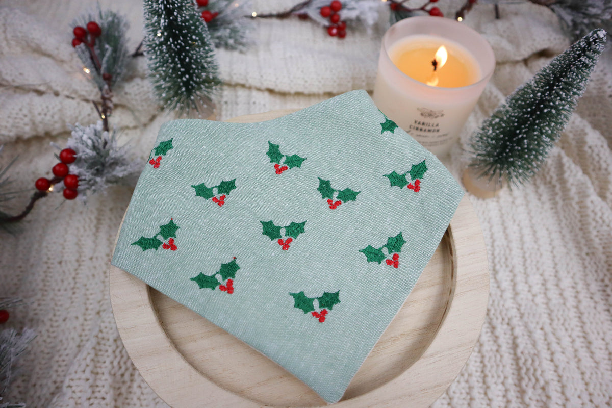 green dog bandana with holly pattern on a wooden plate, surrounded by Christmas decorations.