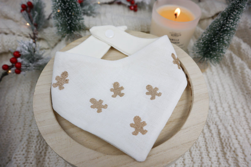 White dog bandana with tan gingerbread man designs on a wooden plate with a candle and Christmas decorations in the background.