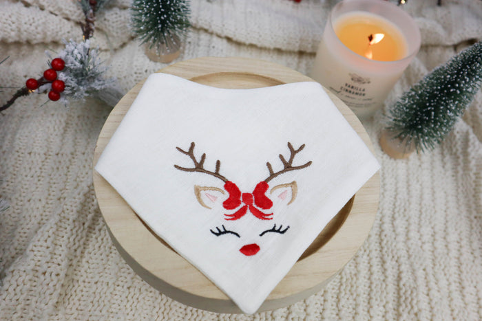 Embroidered scarf with a reindeer face design on a wooden coaster, surrounded by festive decorations.