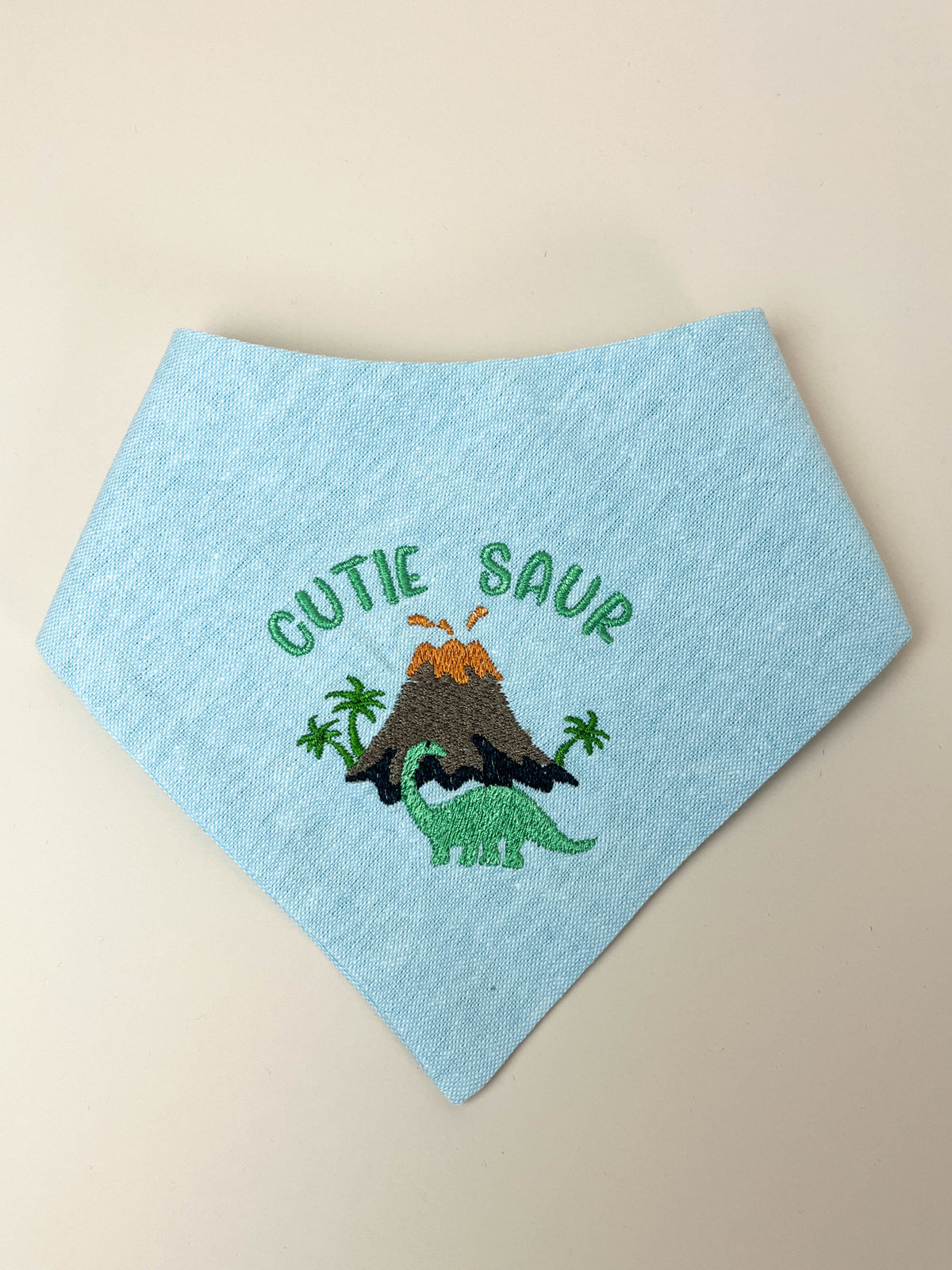 Light blue bandana with dinosaur design and 'Cutie Saur' text on a beige background
