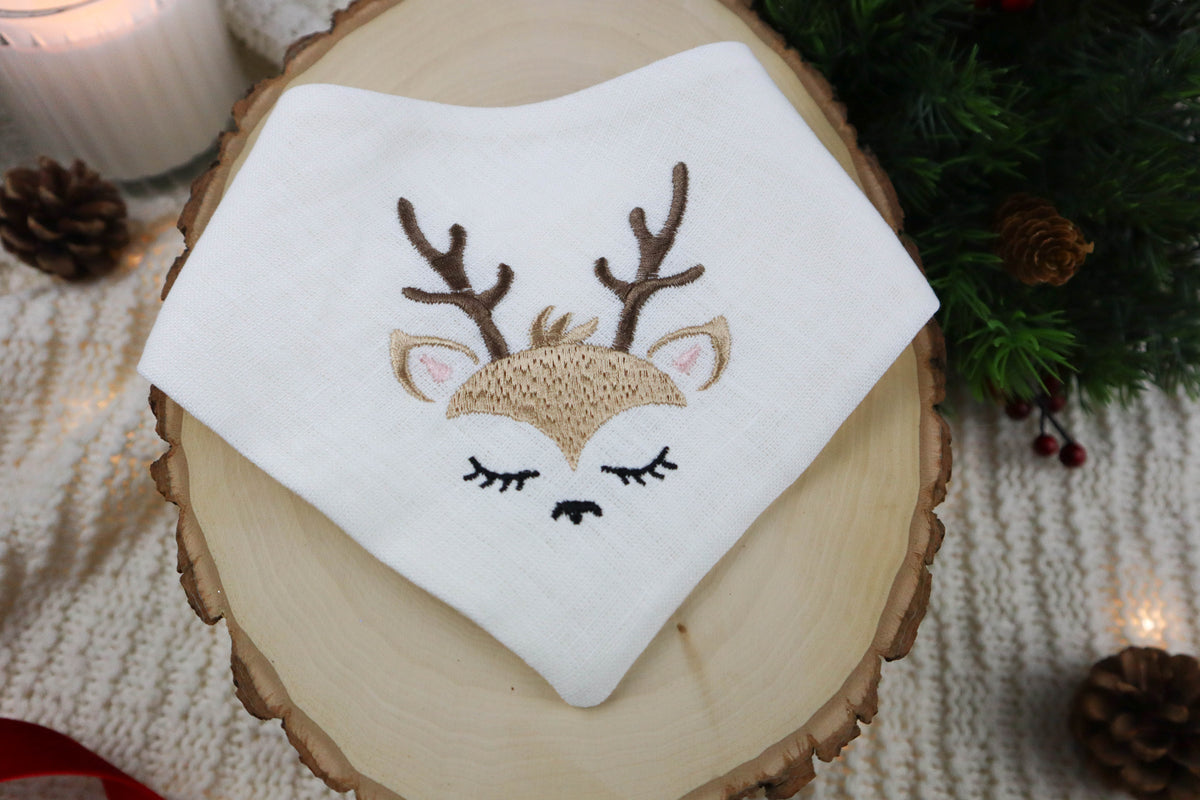 Embroidered deer face on a white dog scarf, placed on a wooden surface with Christmas decorations.