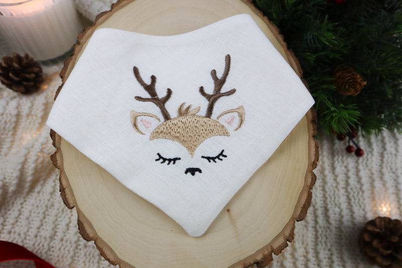 Embroidered deer face on a white dog scarf, placed on a wooden surface with Christmas decorations.