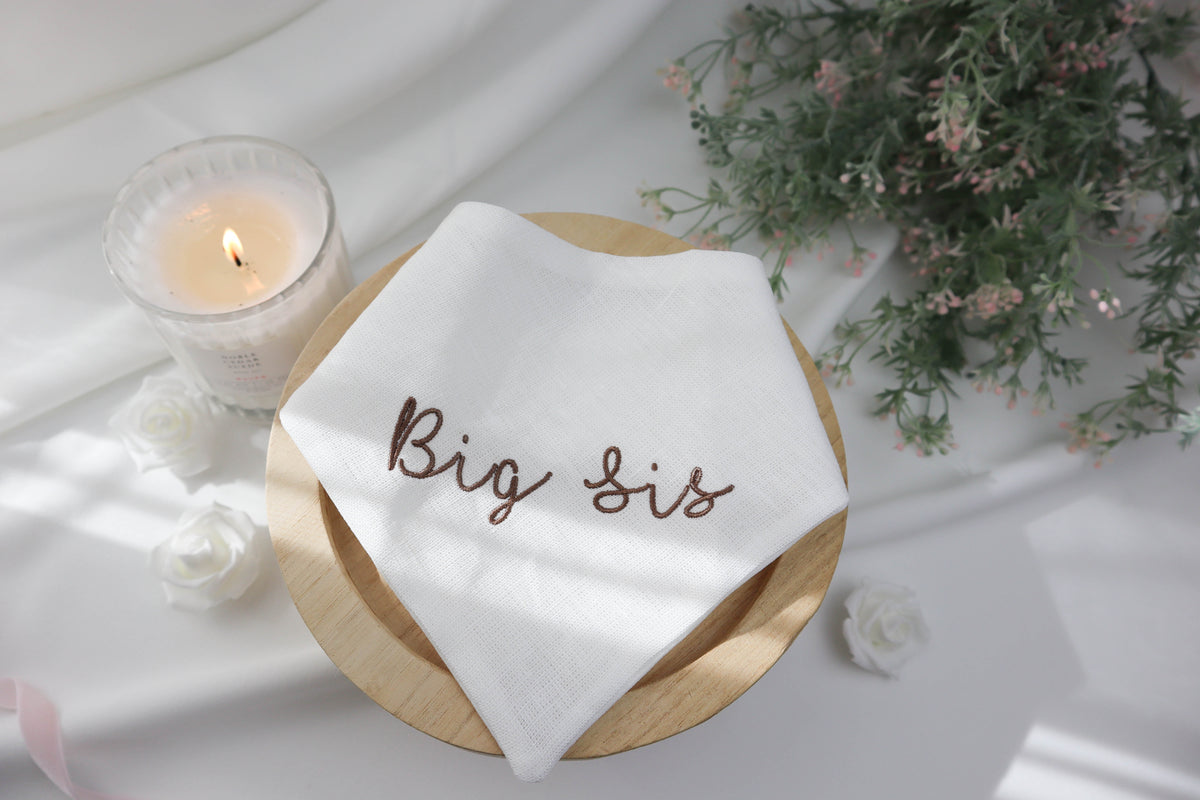 White dog scarf with 'Big Sis' embroidery on a wooden plate with a candle and flowers in the background.