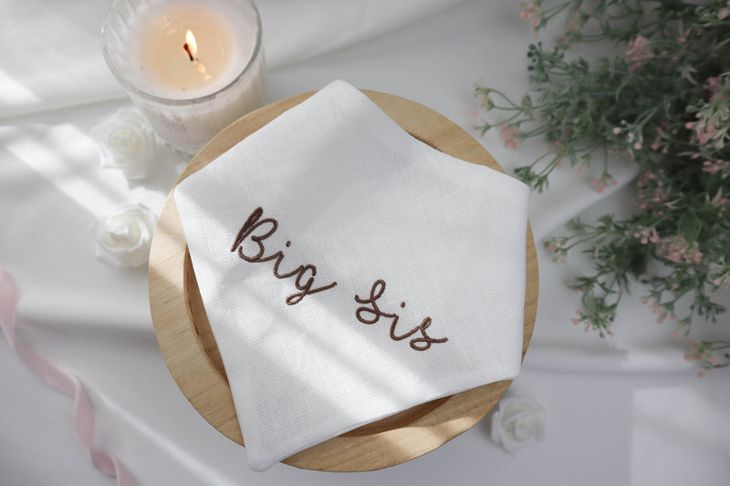 White cat scarf with 'Big Sis' embroidery on a wooden plate with a candle and flowers in the background.