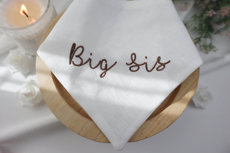 White cat bandana with 'Big Sis' embroidery on a wooden block, lit candle in the background