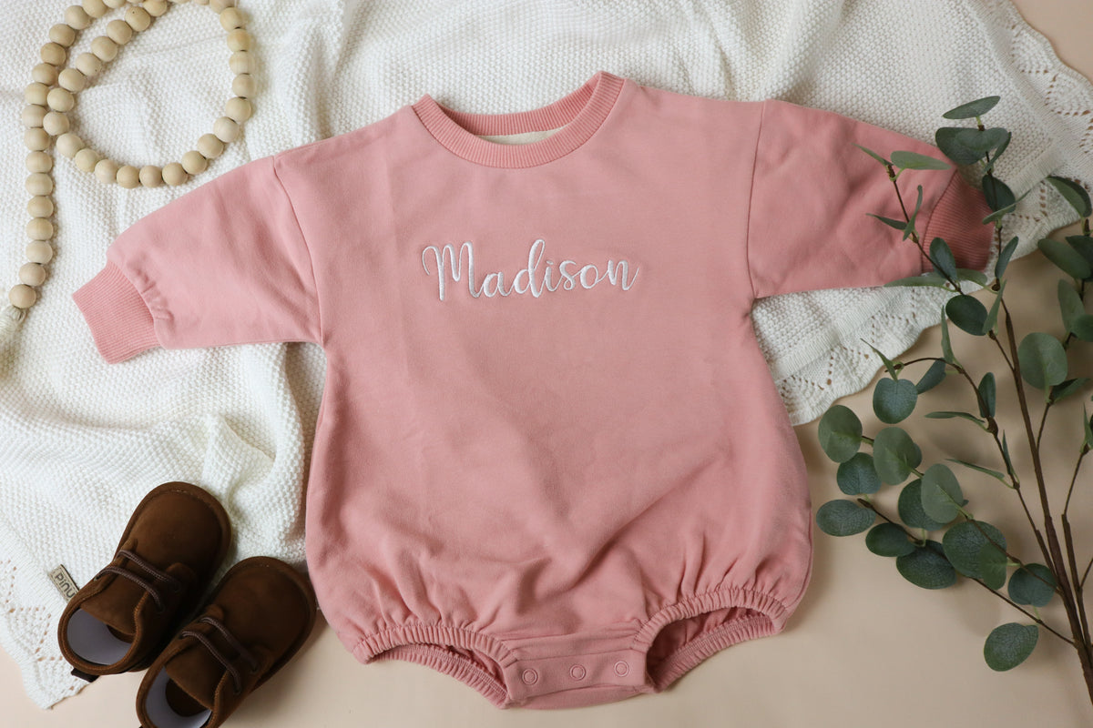 Pink baby romper with 'Madison' embroidered on it, placed on a textured surface with a plant and shoes.