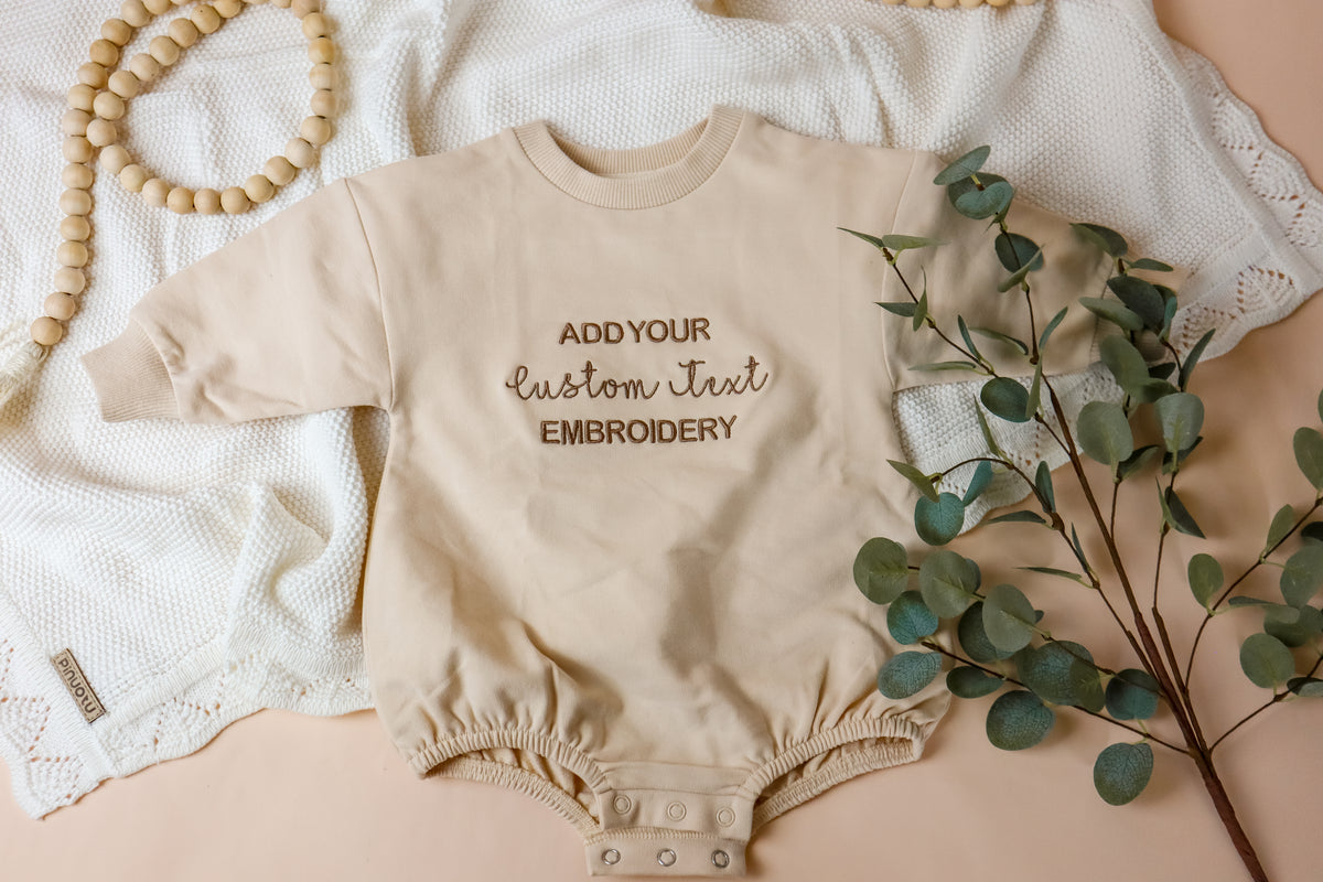 Beige baby romper with text embroidery on a neutral background with eucalyptus leaves.