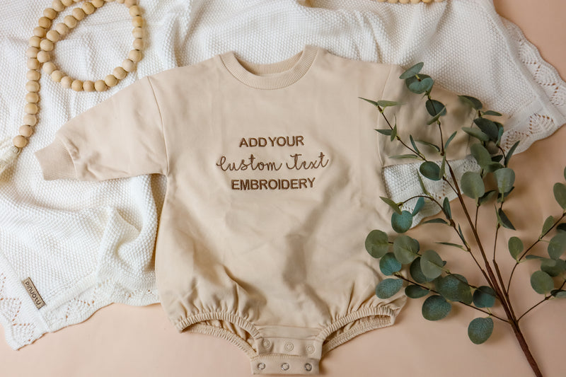 Beige baby romper with text embroidery on a neutral background with eucalyptus leaves.