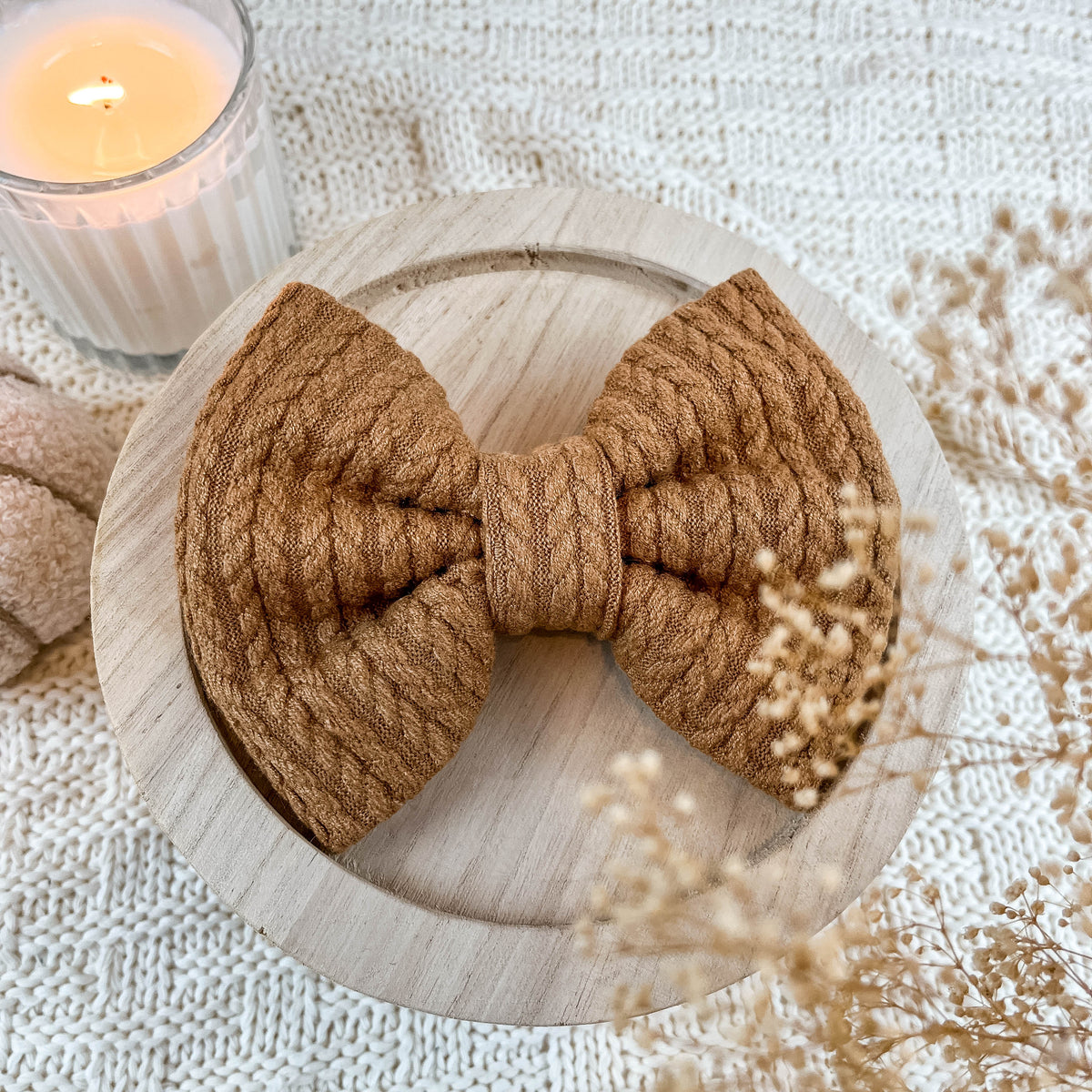 Brown knitted bow on a wooden stand with a candle and dried flowers in the background