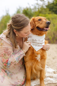 Woman holding a dog with a pregnancy announcement bandana outdoors.