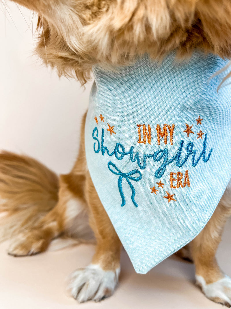 Dog wearing a light blue bandana with 'In My Showgirl Era' text on a plain background