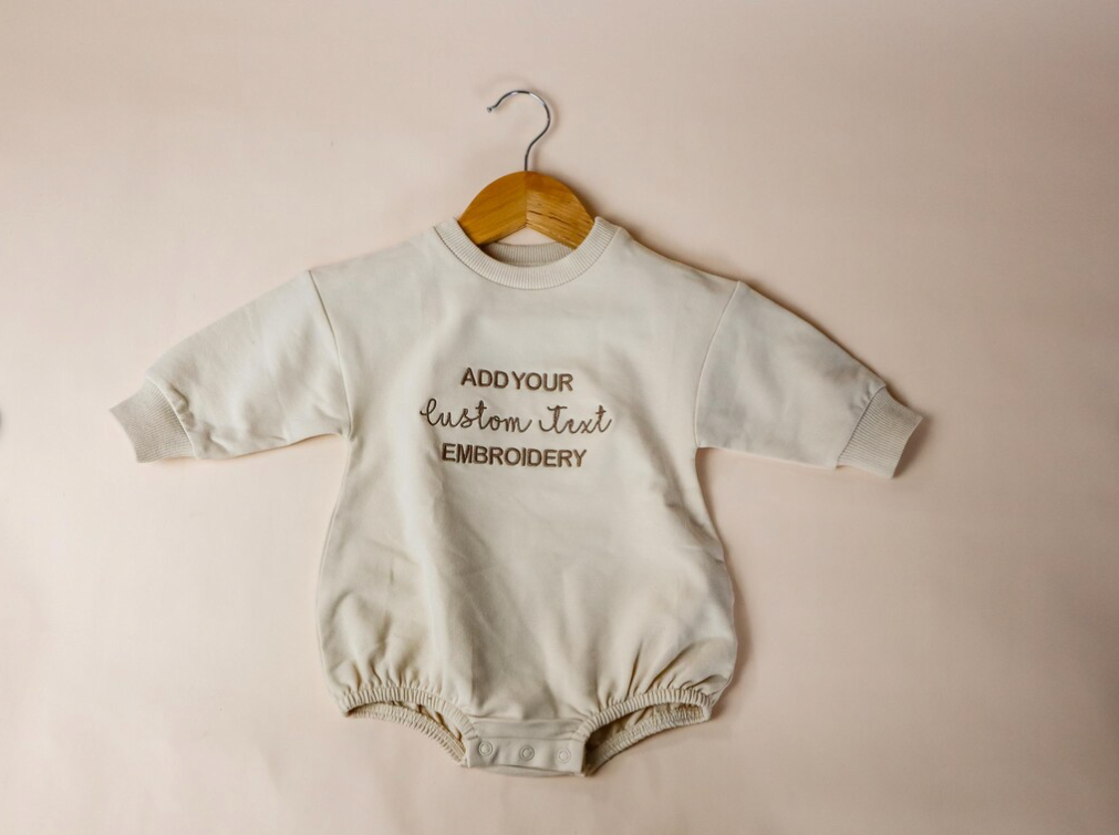 custom embroidered Baby romper with text on a hanger against a beige background
