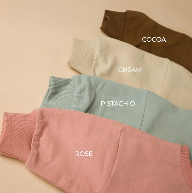 Four different colored baby rompers labeled 'Cocoa', 'Cream', 'Pistachio', and 'Rose' on a beige background.