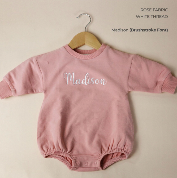 Pink baby romper with 'Madison' embroidered on it, hanging on a wooden hanger against a beige background.