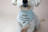 White dog wearing a light blue bandana with 'Baby Johnson Coming Soon' text on a plain background