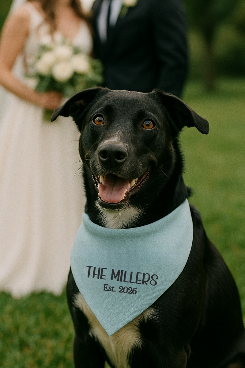 Black dog wearing a light blue bandana with 'The Millers Est. 2026' text, standing in front of a blurred wedding couple.